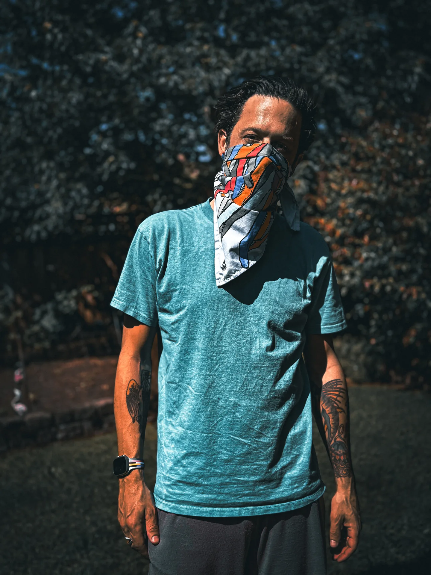 Model in a teal tee with a bright Deafiant bandana worn over the face, standing in a sunny backyard.