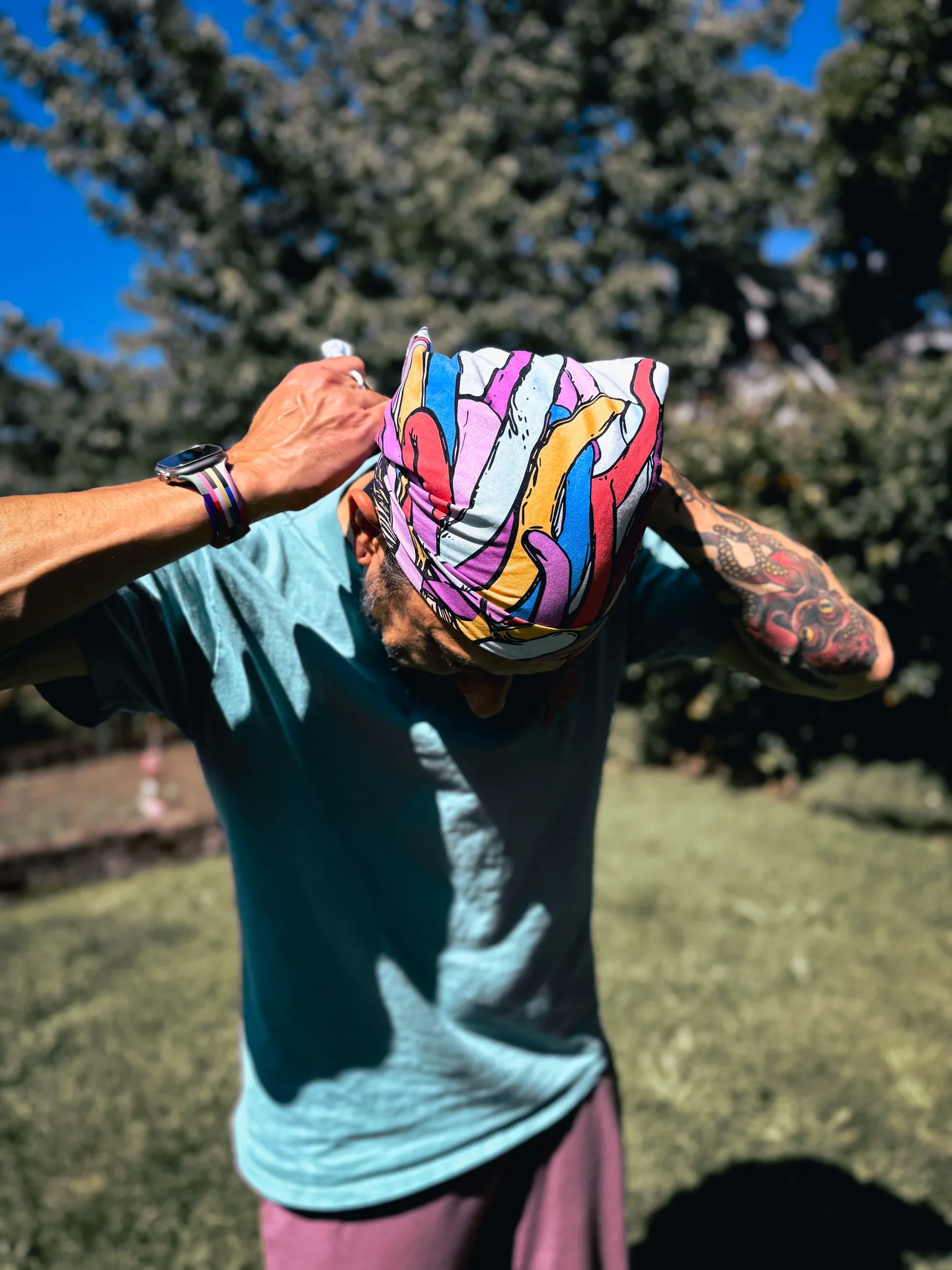 Model tying a bright Deafiant bandana over their head under a blue summer sky.