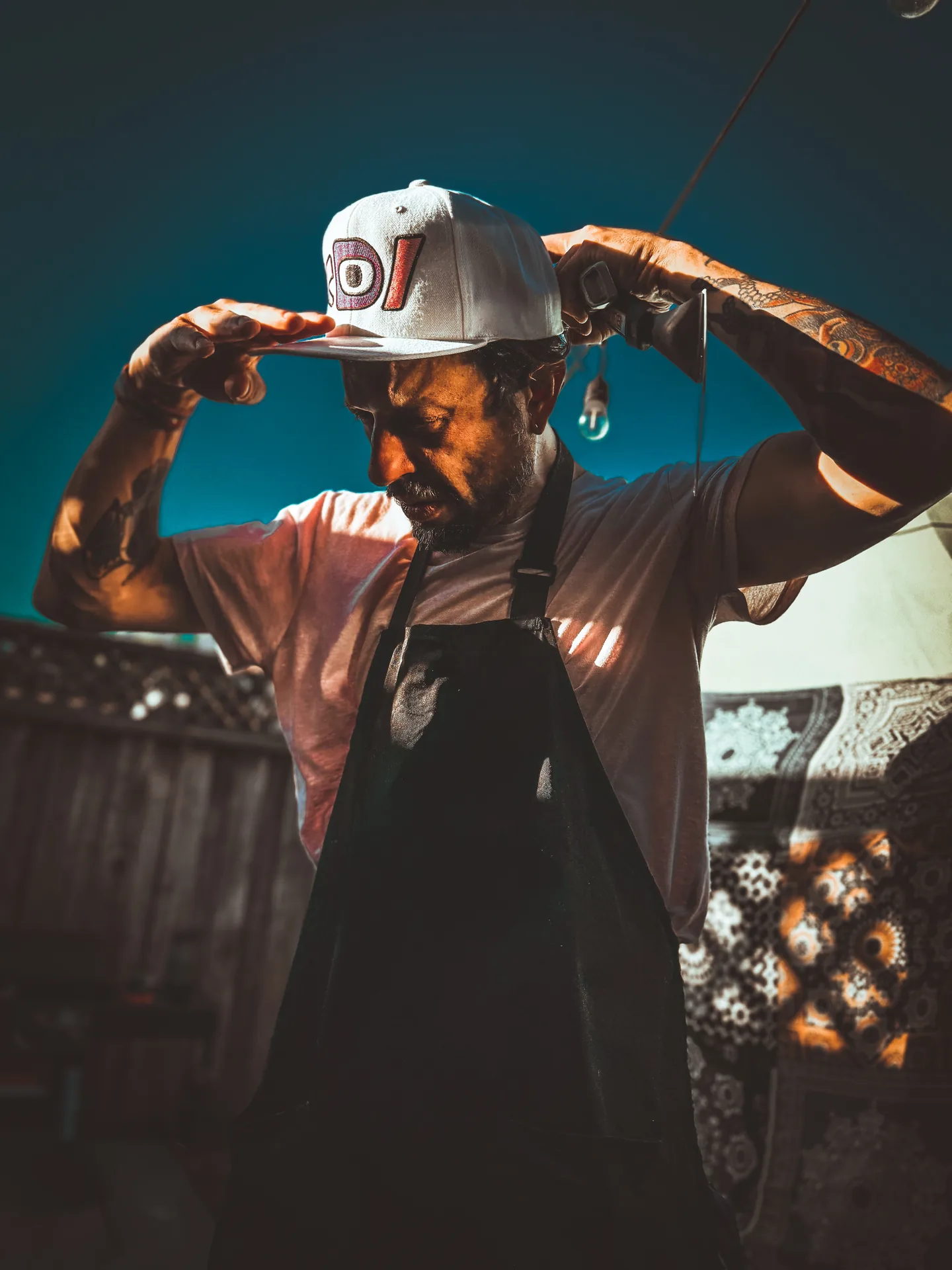 Model in an apron adjusting a white Deafiant snapback with both hands in warm evening light.