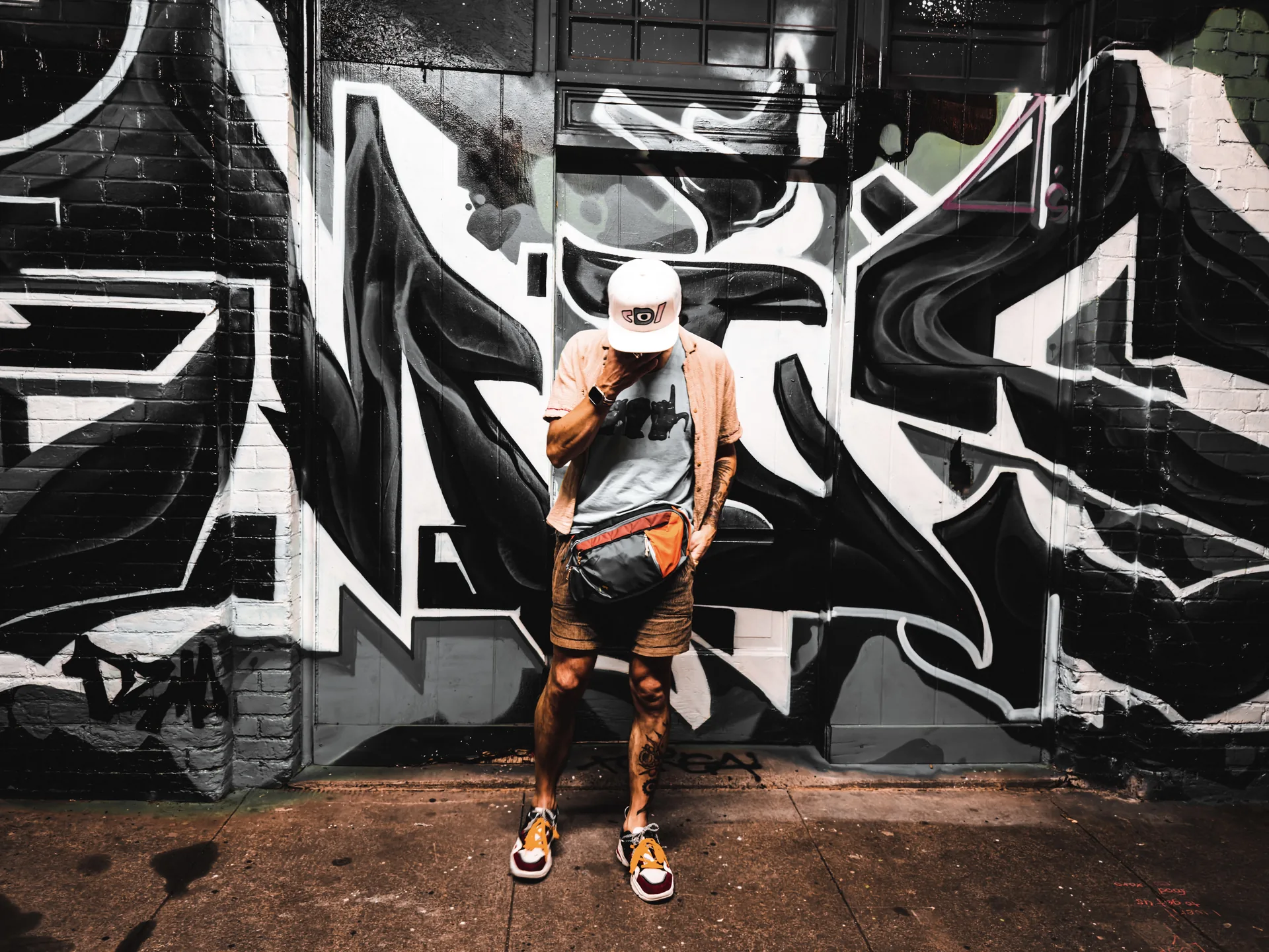 Model in a white Deafiant snapback and sling bag standing in front of a black-and-white graffiti mural at night.