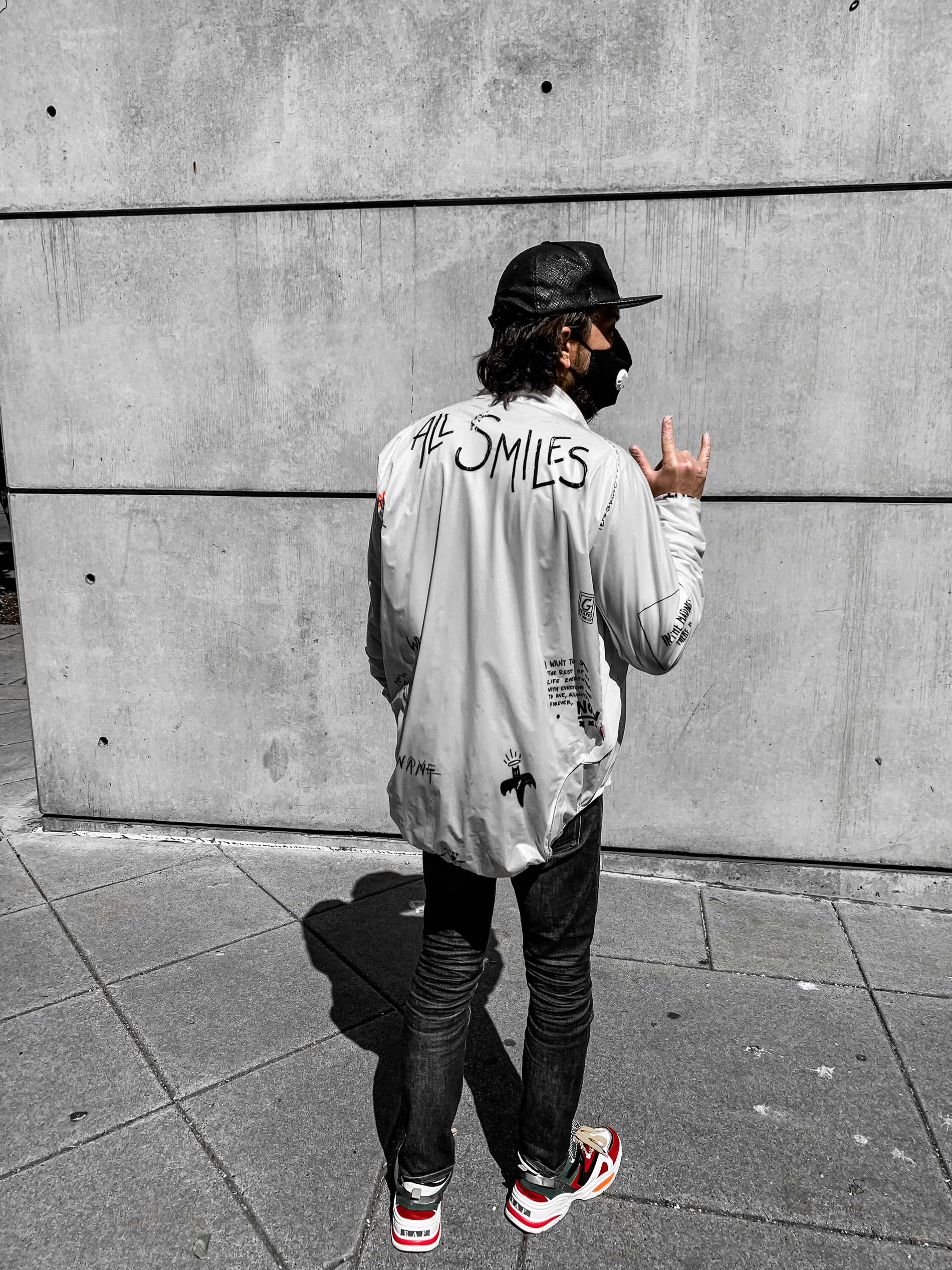 JR in a hand-painted ALL SMILES jacket throwing the ILY hand sign against a concrete wall in Portland. Selective-color treatment with red sneakers as the only color in the frame.